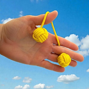 May include: Bright yellow Kendama balls, each intricately knotted, connected by a yellow cord. The balls are held against a blue sky background with fluffy white clouds. The toy is designed for hand-eye coordination.