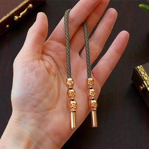 Pode incluir: Duas cordas de cordão trançado verde e preto com contas de caveira douradas e peças finais cilíndricas. O cordão é segurado em uma mão, com um fundo marrom escuro e uma pequena planta em vaso.