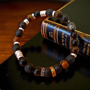 Può includere: Un braccialetto di perline con perline marrone scuro, nere e occhio di tigre. Il braccialetto include distanziatori color argento e un piccolo ciondolo a forma di testa di Buddha. Le perline sono infilate, creando un accessorio elegante.