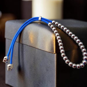Op de afbeelding: Een blauwe koordarmband met zilveren kralen en schedelbedels. De armband heeft een rij kleine, glanzende zilveren kralen en twee blauwe koorden die eindigen in kleine schedelbedels. Het ligt op een donkergrijze doos.