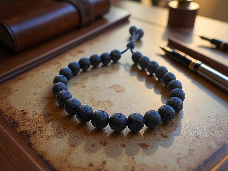 May include: A dark gray beaded bracelet made of porous, round stones. The bracelet is strung on a dark blue cord and rests on a textured, light brown surface. Other items, including a leather-bound notebook and pens, are in the background.