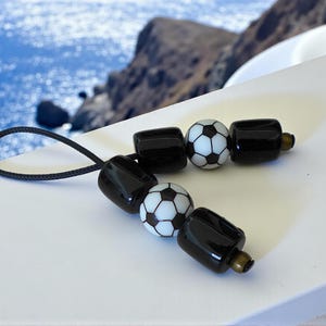 Può includere: Un portachiavi con perline bianche e nere con due perline a forma di pallone da calcio e quattro perline rettangolari nere. Il portachiavi ha un anello di corda nero. Lo sfondo è un'immagine sfocata del mare e di una costa rocciosa.