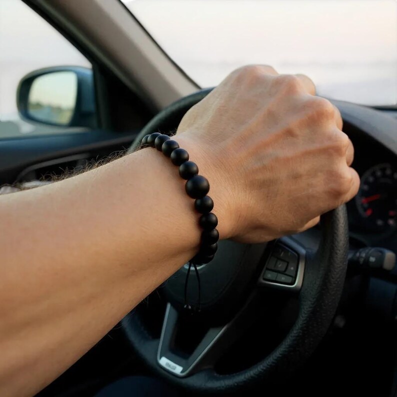 May include: A black beaded bracelet with a matte finish. The bracelet is made of round beads of varying sizes and is worn on the wrist. The bracelet is strung on a black cord.