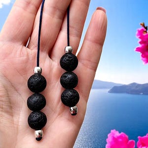 Peut inclure: Boucles d'oreilles en perles de roche volcanique noire. Chaque boucle d'oreille comporte trois perles noires rondes et texturées, séparées par des perles métalliques argentées. Les boucles sont suspendues à un cordon bleu foncé.