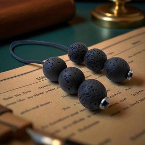 May include: Six dark grey lava stone beads with a textured surface are strung on a grey cord. The beads are arranged on vintage paper with text. The beads have a silver-coloured metal screw on the end.