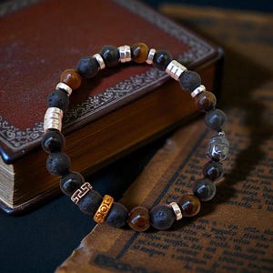 May include: A bracelet made with black lava, tiger's eye, and silver beads. It features a silver Buddha charm and gold spacers. The bracelet is displayed on a dark surface next to a vintage book, with the beads measuring in cm.