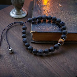 May include: Black beaded bracelet featuring matte onyx and lava stone beads, with a single tiger eye bead and silver-tone accents. The bracelet is adjustable and rests on a dark wooden surface.