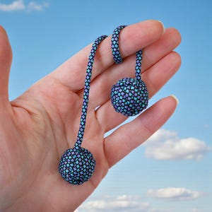 Puede incluir: Primer plano de una mano sosteniendo un par de begleri. Los begleri están hechos de cordón tejido en tonos de azul, morado y negro, con forma esférica en cada extremo. El fondo es un cielo azul brillante con nubes blancas y esponjosas.
