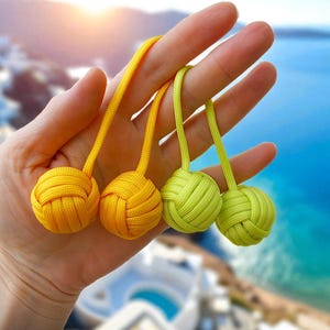 May include: Four decorative knot balls in shades of yellow and green are held in a hand. The balls are made of woven cord and have a loop for hanging. The background shows a blurred view of the ocean and buildings.
