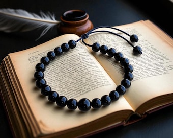 May include: A black beaded bracelet rests on an open antique book with aged pages. The bracelet is made of smooth, round beads and is strung on a dark cord. A feather and inkwell are in the background, creating a vintage aesthetic.