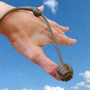 Könnte beinhalten: Ein geknotetes Seilspielzeug mit einer Kugel am Ende, in Grün-, Braun- und Orangetönen. Das Seil ist zu einem Armband geschlungen. Das Spielzeug wird vor einem blauen Himmelshintergrund gehalten.