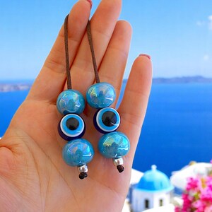 Puede incluir: Dos conjuntos de abalorios de cuentas de vidrio azul con un cordón marrón. Cada conjunto tiene tres cuentas, dos azules iridiscentes y una con un diseño de ojo. El fondo muestra un mar y un cielo azules.