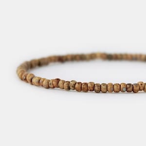 Könnte beinhalten: Ein einreihiges Armband aus kleinen, runden Perlen in Erdtönen. Die Perlen haben eine gesprenkelte, texturierte Oberfläche mit Variationen in Beige-, Braun- und gedeckten Grautönen, die in einer durchgehenden kreisförmigen Schleife angeordnet sind.