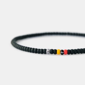 Puede incluir: Un brazalete de cuentas negras con una sola cuenta blanca, negra y amarilla, seguida de una cuenta roja.
