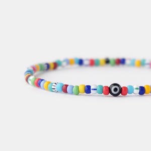 May include: A colourful beaded bracelet featuring a blue and white evil eye charm. The bracelet is made with small, round beads in a variety of colours, including red, yellow, blue, green, and white.