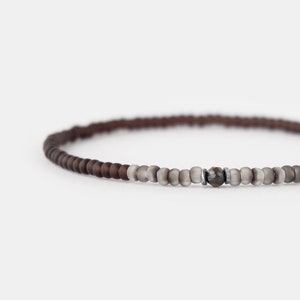 Könnte beinhalten: Ein braunes und graues Perlenarmband mit einer silbernen Perle in der Mitte.