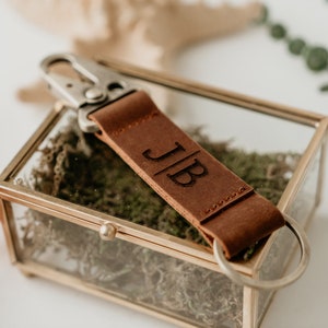 May include: A brown leather keychain with a silver metal clasp and a ring. The keychain is personalized with the initials "JB".