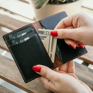 Może przedstawiać: Brązowy sk&oacute;rzany portfel z klapką, otwarty, aby pokazać dwie karty kredytowe i banknot dwudziestodolarowy. Portfel ma obszyty brzeg i brązowe wnętrze.
