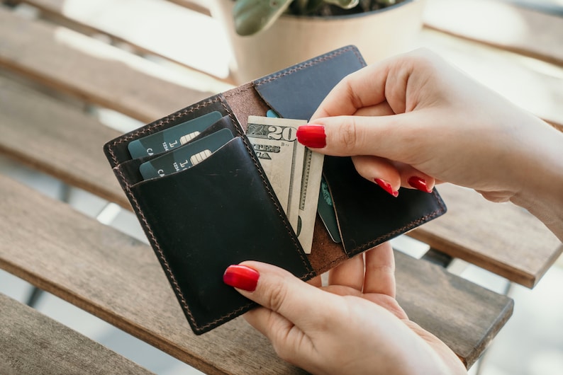 Może przedstawiać: Brązowy sk&oacute;rzany portfel z klapką, otwarty, aby pokazać dwie karty kredytowe i banknot dwudziestodolarowy. Portfel ma obszyty brzeg i brązowe wnętrze.