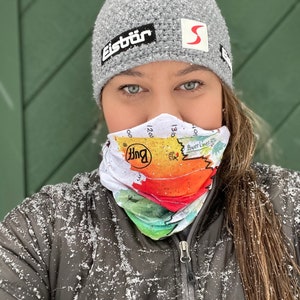 May include: A woman wearing a gray knit hat with a red pom pom and the word "Eistür" on it. She is also wearing a colorful neck gaiter with the word "Buff" on it. The woman is standing in front of a green background and there is snow falling around her.