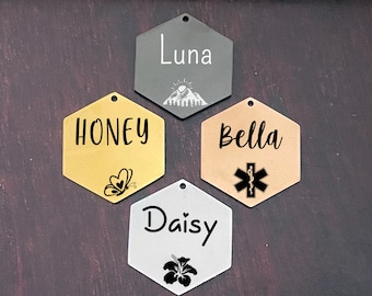 May include: Four hexagonal pet ID tags with different designs. The tags are made of metal and have a hole at the top for attaching to a collar. The tags are engraved with the names "Luna", "Honey", "Bella", and "Daisy". Each tag has a unique design, including a mountain, a butterfly, a medical symbol, and a flower.