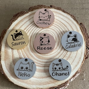 May include: Five round cat-themed pet ID tags with different names engraved on them. The tags are in gold, silver, and rose gold colours. The names on the tags are Cashew, Sauron, Reese, Galadriel, and Neko.