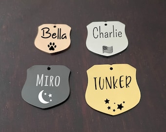May include: Four personalised pet ID tags in the shape of shields. The tags are made of metal and have different colours and designs. The names "Bella", "Charlie", "Miro", and "Tunker" are engraved on the tags. Bella's tag is rose gold with a paw print, Charlie's tag is silver with an American flag, Miro's tag is black with a crescent moon and stars, and Tunker's tag is gold with stars.