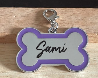 Puede incluir: Una placa de identificación para perro en forma de hueso, de color morado y plateado, con el nombre "Sami" grabado en ella.