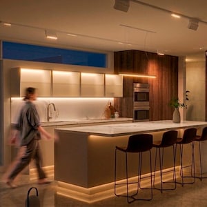 May include: A modern kitchen featuring a large white marble island with four dark brown bar stools, under-cabinet lighting, and tall dark wood cabinetry. The space includes a stainless steel refrigerator, built-in ovens, and track lighting.