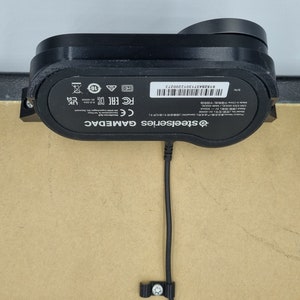 May include: Black SteelSeries GAMEDAC external sound card with a black cable and a black clip. The device has a label with the text "SteelSeries GAMEDAC" and "CE FC EACC" and a serial number.