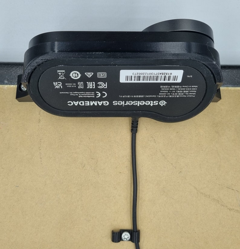 May include: Black SteelSeries GAMEDAC external sound card with a black cable and a black clip. The device has a label with the text "SteelSeries GAMEDAC" and "CE FC EACC" and a serial number.
