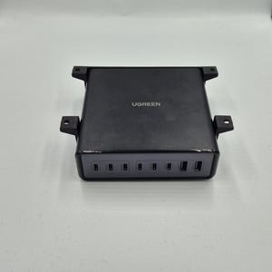 Op de afbeelding: Een zwarte UGREEN-oplaadstation met meerdere poorten. Het apparaat heeft een rechthoekige vorm met de merknaam erop gedrukt. Het beschikt over verschillende USB-poorten en een strak, modern ontwerp.