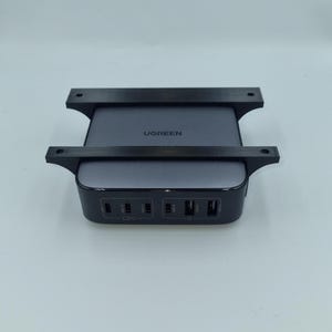 May include: A dark grey UGREEN charging station with multiple ports, secured by a black bracket. The charging station has the UGREEN logo. The black bracket has a distinctive design, holding the charger securely.