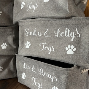 May include: Three gray fabric storage bins with white paw prints and the names "Rune's Toys", "Simba & Lolly's Toys", and "Leo & Roxy's Toys" printed on them.
