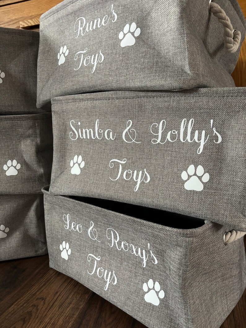 May include: Three gray fabric storage bins with white paw prints and the names "Rune's Toys", "Simba & Lolly's Toys", and "Leo & Roxy's Toys" printed on them.