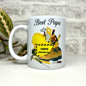 May include: White ceramic mug with a black handle. The mug features a graphic of a yellow hard hat, tools, and work boots with the text "Best Papa" and "Under Construction".