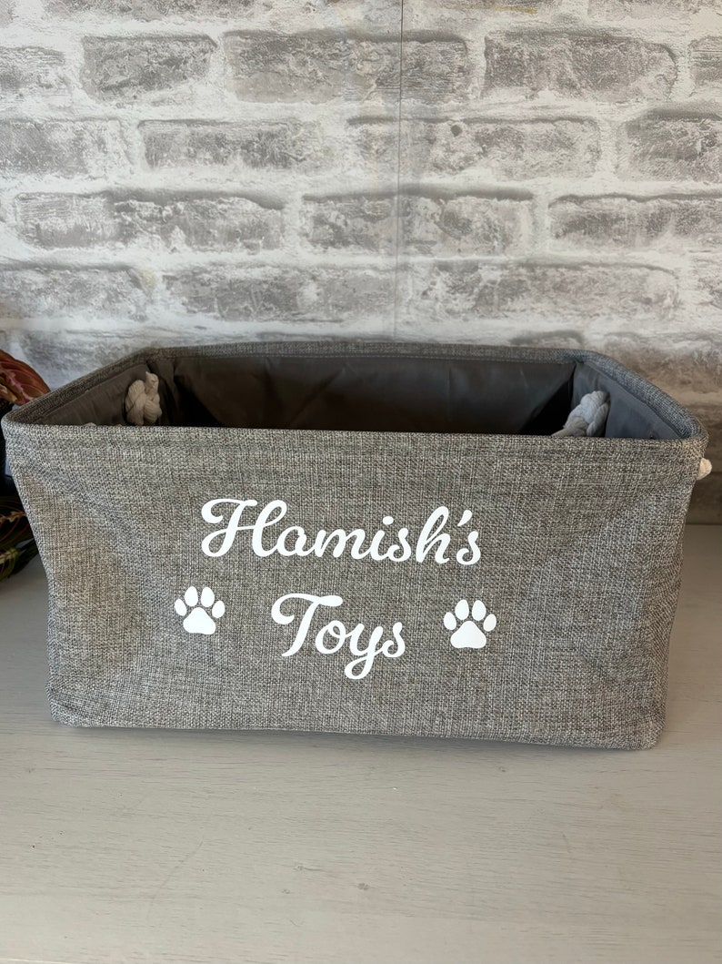 May include: A gray fabric storage bin with the text "Hamish's Toys" and two white paw prints embroidered on the front.