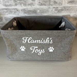 May include: A gray fabric storage bin with the text "Hamish's Toys" and two white paw prints.