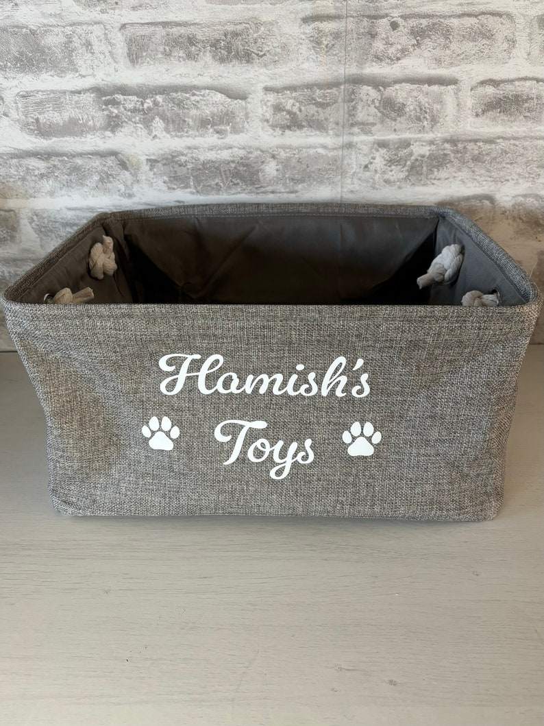 May include: A gray fabric storage bin with the text "Hamish's Toys" and two white paw prints.