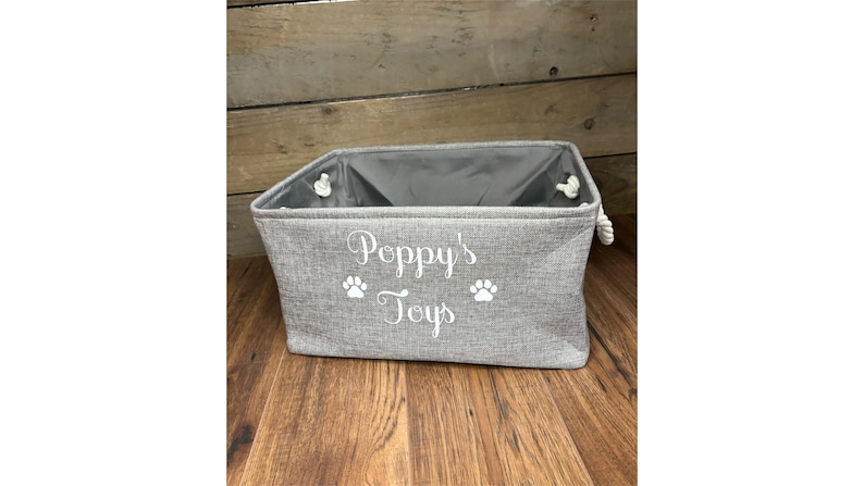 May include: A gray fabric storage bin with white rope handles. The bin has the text "Poppy's Toys" and two paw prints embroidered on the front.