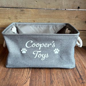 May include: A gray canvas storage bin with white rope handles. The bin has the text "Cooper's Toys" and two paw prints printed on the front.