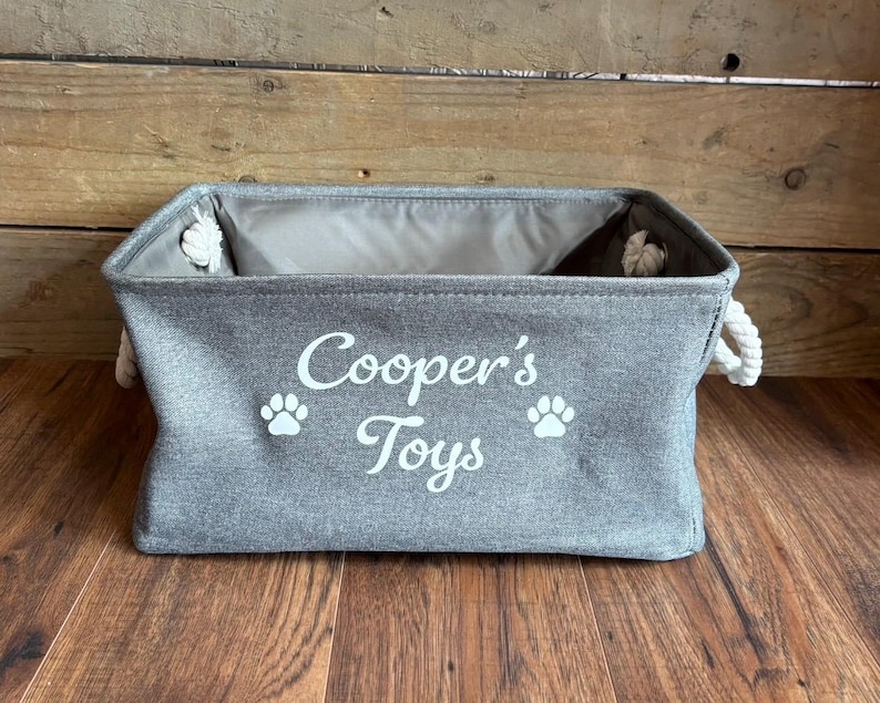 May include: A gray canvas storage bin with white rope handles. The bin has the text "Cooper's Toys" and two paw prints printed on the front.