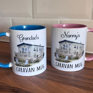 May include: Two white ceramic mugs featuring a caravan illustration. The left mug has a blue handle and interior with the text Grandad's CARAVAN MUG. The right mug has a pink handle and interior with the text Nanny's CARAVAN MUG printed above the caravan.