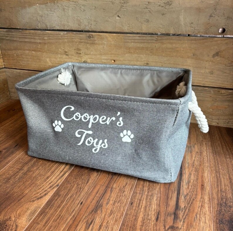 May include: A gray canvas storage bin with rope handles. The bin has white paw prints and the text "Cooper's Toys" printed on the front.
