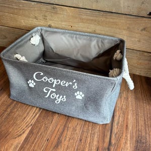 May include: A gray canvas storage bin with rope handles. The bin has white paw prints and the text "Cooper's Toys" printed on the front.