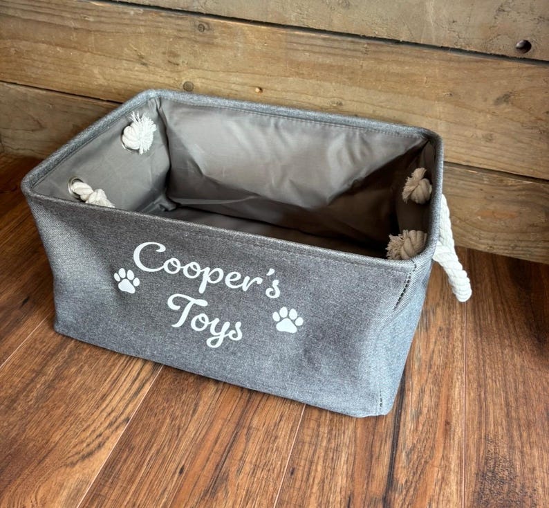 May include: A gray canvas storage bin with rope handles. The bin has white paw prints and the text "Cooper's Toys" printed on the front.