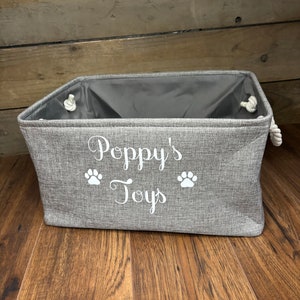 May include: A gray fabric storage bin with white rope handles. The bin has the text "Poppy's Toys" and two white paw prints on the front.