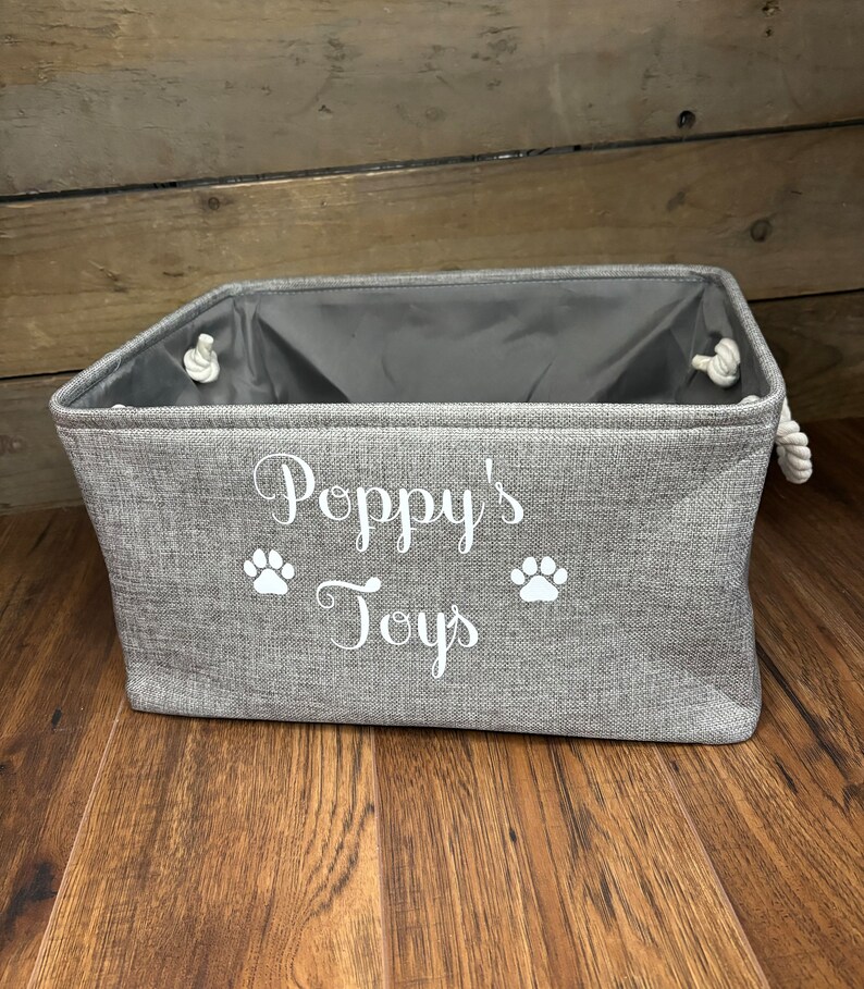 May include: A gray fabric storage bin with white rope handles. The bin has the text "Poppy's Toys" and two white paw prints on the front.