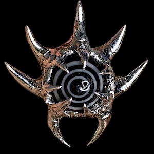 May include: A metallic silver brooch featuring a textured, spiked outer frame surrounding a circular black and white target pattern. The centre of the design displays a small, white, stylised letter D symbol set against the concentric rings.