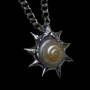 Puede incluir: Un collar de plata con un colgante de sol espinoso con una concha en el centro.